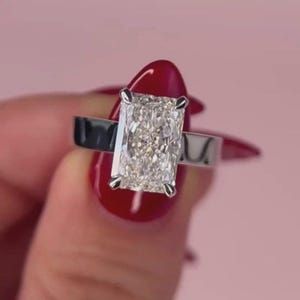 Op de afbeelding: Een ring met een radiant-geslepen diamant, voorzien van een grote, rechthoekige, gefacetteerde heldere steen in een gepolijste zilverkleurige metalen band. De centrale steen vertoont meerdere facetten die licht reflecteren, vastgezet met vier pootjes.