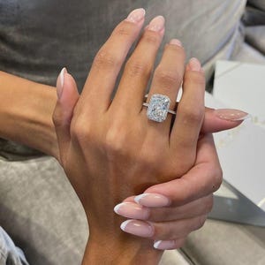 Op de afbeelding: Een verlovingsring met een radiant-geslepen diamant, voorzien van een grote rechthoekige centrale steen omringd door een halo van kleinere pavé-diamanten, geplaatst op een dunne, verfijnde metalen band bezet met kleine diamanten langs de ring.