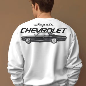Könnte beinhalten: Ein weißes Langarm-Sweatshirt mit einem schwarzen Grafikdruck auf der Rückseite. Das Design zeigt das Wort Impala in Schreibschrift, das Wort CHEVROLET in fetten Großbuchstaben und eine detaillierte Seitenansicht eines schwarzen Oldtimers.