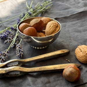 Peut inclure: Un casse-noix en métal argenté placé à côté d'un petit bol en argent rempli de noix entières, incluant des noix et des noisettes. Une noisette et une noix isolées reposent à proximité, accompagnées de brins de lavande séchée violette.