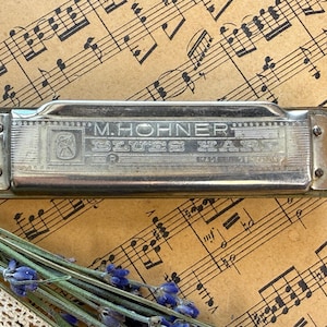 Pode incluir: Uma harmónica M. Hohner Blues Harp de tom prateado com acabamento metálico, apresentando uma gravação com o texto M. HOHNER BLUES HARP e MADE IN GERMANY, fixada com pequenos parafusos em cada lado, com forma retangular e textura metálica.