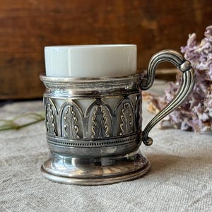 Könnte beinhalten: Eine weiße Porzellantasse in einem dekorativen silberfarbenen Metallhalter. Der Halter zeichnet sich durch ein komplexes durchbrochenes Design mit vertikalen blattartigen Motiven und einem geschwungenen Griff aus. Das Metall ist poliert.