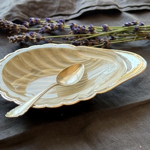 May include: A shell-shaped dish with a glossy, cream-colored interior featuring subtle gray marbling and a scalloped silver-toned rim. A small, polished silver-toned spoon rests inside the dish, which is positioned near a sprig of dried purple lavender.