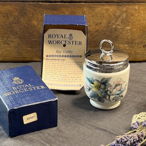 May include: A Royal Worcester egg coddler with a floral pattern, featuring a white porcelain base and a silver-tone metal lid with a ring handle. The item is displayed with its original blue cardboard box, which is labelled with the name Astley.