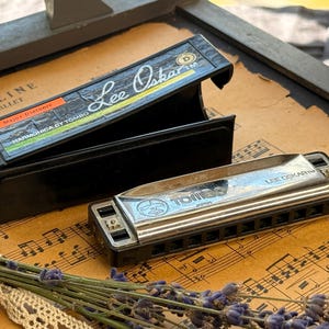Könnte beinhalten: Eine silberne Lee Oskar Major Diatonic Mundharmonika von Tombo mit schwarzem Kunststoffkamm und Metallabdeckungen, begleitet von ihrem schwarzen Kunststoffetui. Das Etui trägt ein Etikett mit dem Text Lee Oskar, Major Diatonic und Harmonica by Tombo.