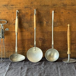 Pode incluir: Uma coleção de cinco utensílios de cozinha vintage, incluindo um batedor de ovos rotativo de metal, uma concha com cabo de madeira, uma escumadeira perfurada com cabo de madeira, uma concha de metal e um espremedor de batatas com cabo de madeira.
