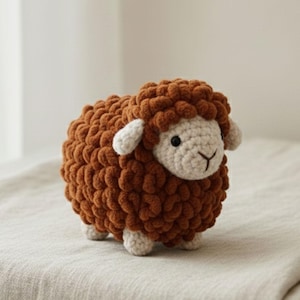 May include: A small, handmade crochet sheep plush toy featuring a textured, dark brown woolly body and a cream-colored face. The sheep has two small cream-colored ears, two black bead eyes, and a simple embroidered black nose and mouth detail.
