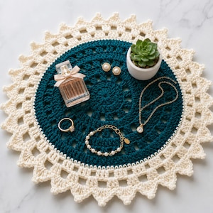 May include: A round, dark teal crochet doily with a cream-colored scalloped border. Arranged on top are a glass perfume bottle with a beige bow, a small white pot containing a green succulent, two pearl earrings, a gold ring, and a gold necklace.