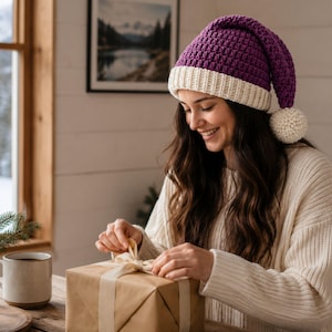 May include: A purple crocheted hat with a cream-colored ribbed brim and a matching cream pom-pom at the tip. The hat features a textured stitch pattern and is worn with a cream-colored knit sweater while wrapping a brown paper gift box.