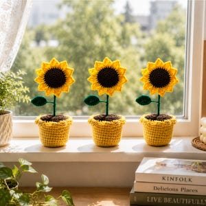 May include: Three crocheted sunflowers in yellow pots. Each flower has a dark brown center, bright yellow petals, a green stem with one leaf, and is planted in a textured yellow pot. The sunflowers are arranged in a row on a window ledge.