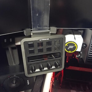 May include: A dark gray Elgato Stream Deck Plus control console featuring eight programmable LCD keys, a central touch strip, and four rotary dials. The device is mounted to a monitor using a clear plastic bracket and displays the text STREAM DECK.