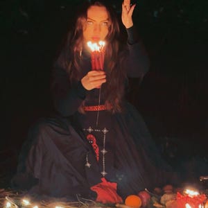 Peut inclure: Une robe noire &agrave; manches longues avec un motif brod&eacute; rouge sur le corsage et la jupe, port&eacute;e en tenant un bouquet de bougies rouges allum&eacute;es. La sc&egrave;ne comprend de nombreuses petites bougies allum&eacute;es plac&eacute;es au sol devant la robe.