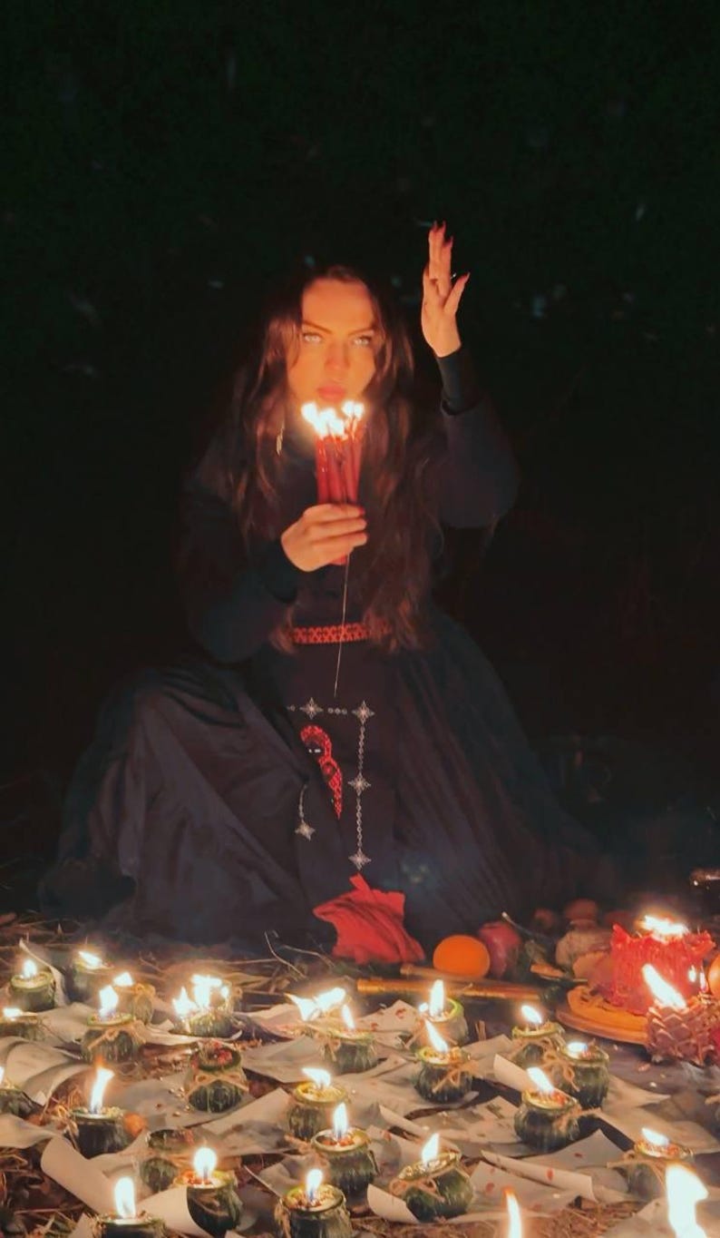 Peut inclure: Une robe noire &agrave; manches longues avec un motif brod&eacute; rouge sur le corsage et la jupe, port&eacute;e en tenant un bouquet de bougies rouges allum&eacute;es. La sc&egrave;ne comprend de nombreuses petites bougies allum&eacute;es plac&eacute;es au sol devant la robe.