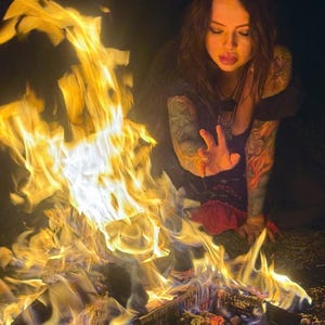 Peut inclure: Un feu de camp aux flammes orange et jaune vives br&ucirc;le intens&eacute;ment au-dessus de b&ucirc;ches carbonis&eacute;es sombres. Le feu est situ&eacute; devant une personne aux cheveux fonc&eacute;s et aux bras tatou&eacute;s qui tend une main en direction des flammes ardentes.
