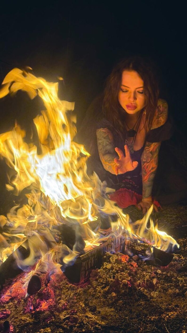 Peut inclure: Un feu de camp aux flammes orange et jaune vives br&ucirc;le intens&eacute;ment au-dessus de b&ucirc;ches carbonis&eacute;es sombres. Le feu est situ&eacute; devant une personne aux cheveux fonc&eacute;s et aux bras tatou&eacute;s qui tend une main en direction des flammes ardentes.