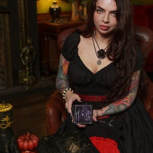 Peut inclure: Un jeu de cartes de tarot noir avec une illustration violette d'une maison et sept lignes verticales, tenu par une personne portant un haut noir &agrave; accents rouges, un collier superpos&eacute; et un bracelet, avec un livre sombre et des citrouilles orange.