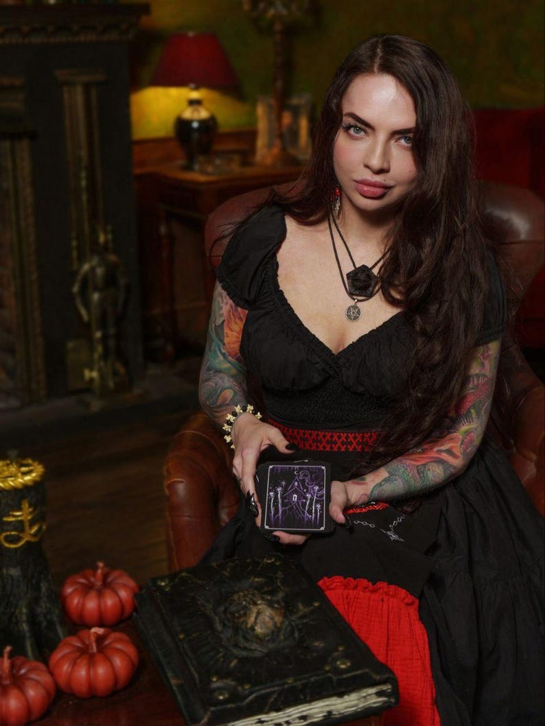 Peut inclure: Un jeu de cartes de tarot noir avec une illustration violette d'une maison et sept lignes verticales, tenu par une personne portant un haut noir &agrave; accents rouges, un collier superpos&eacute; et un bracelet, avec un livre sombre et des citrouilles orange.