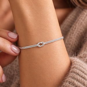 Könnte beinhalten: Ein silberfarbenes Armband mit einem zarten, zweireihigen Kettendesign, das in einem zentralen Knotendetail zusammenläuft. Das Armband ist mit einer glatten, flexiblen Metalloberfläche und einem sicheren Verschlussmechanismus gefertigt.