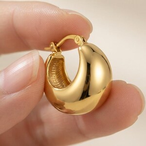 May include: A small, thick, gold-toned hoop earring with a polished, reflective surface. The earring features a hinged closure mechanism at the top, with a smooth, rounded, and chunky design that curves into a teardrop-like shape.