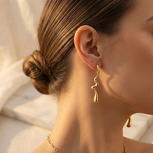 May include: A gold-toned earring featuring a long, slender, and fluid wavy design that hangs vertically from the earlobe. The metal surface is smooth and polished, reflecting light with a bright, metallic sheen along its curved, organic shape.