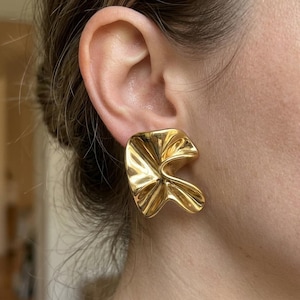 May include: A gold-toned earring with a polished, reflective surface and an irregular, folded, or rippled shape. The earring features a smooth, metallic finish with deep curves and indentations that create a dynamic, sculptural appearance on the earlobe.