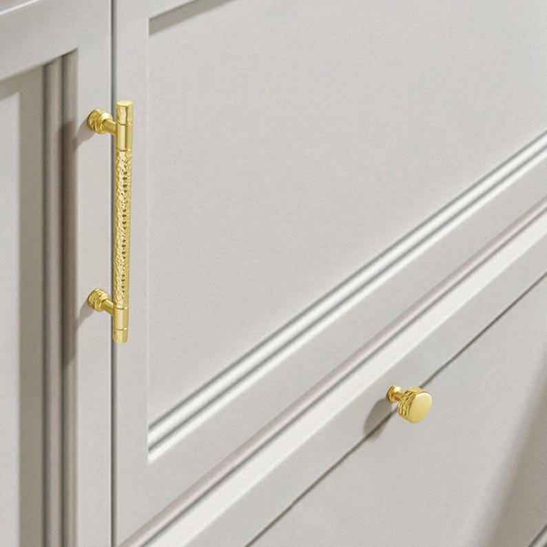 May include: A gold-toned metal cabinet handle with a textured, hammered centre section and smooth ends, mounted vertically on a light grey cabinet door, alongside a matching small, round, textured gold-toned cabinet knob on a drawer front.