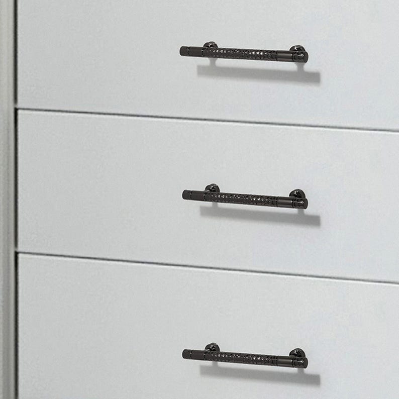 May include: Three matte black drawer pulls with a textured, knurled centre section and smooth ends. Each handle is mounted horizontally onto a flat, light grey surface, featuring two small circular attachment points at the ends of each bar.