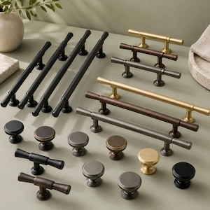 Può includere: Una collezione di ferramenta per mobili in metallo che include lunghe maniglie a barra, maniglie a T e pomelli rotondi in varie finiture come nero opaco, oro spazzolato, bronzo e grigio canna di fucile, disposti su una superficie piana.