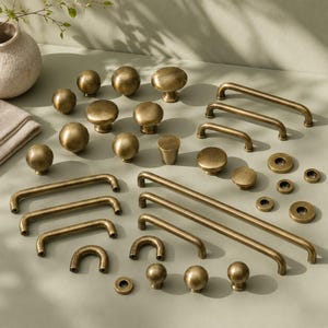 Op de afbeelding: Een collectie kastbeslag van antiek messing, inclusief diverse ronde knoppen, lange stanggrepen, U-vormige handgrepen en kleine ronde ringen. Het beslag is voorzien van een geborstelde metalen afwerking met een warme, gouden tint.