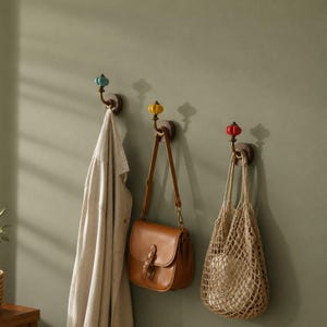 May include: Three wall-mounted hooks with dark wood circular bases and brass-toned metal arms. Each hook features a small, rounded, fluted ceramic knob in light blue, yellow, or red, holding a beige coat, a brown leather bag, and a beige net bag.
