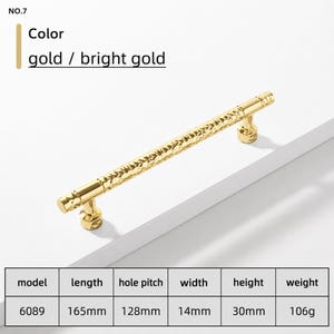 May include: A bright gold cabinet handle with a textured, hammered surface. The handle is labelled No. 7 and model 6089, with a length of 165mm, hole pitch of 128mm, width of 14mm, height of 30mm, and a weight of 106g. The finish is shiny and metallic.