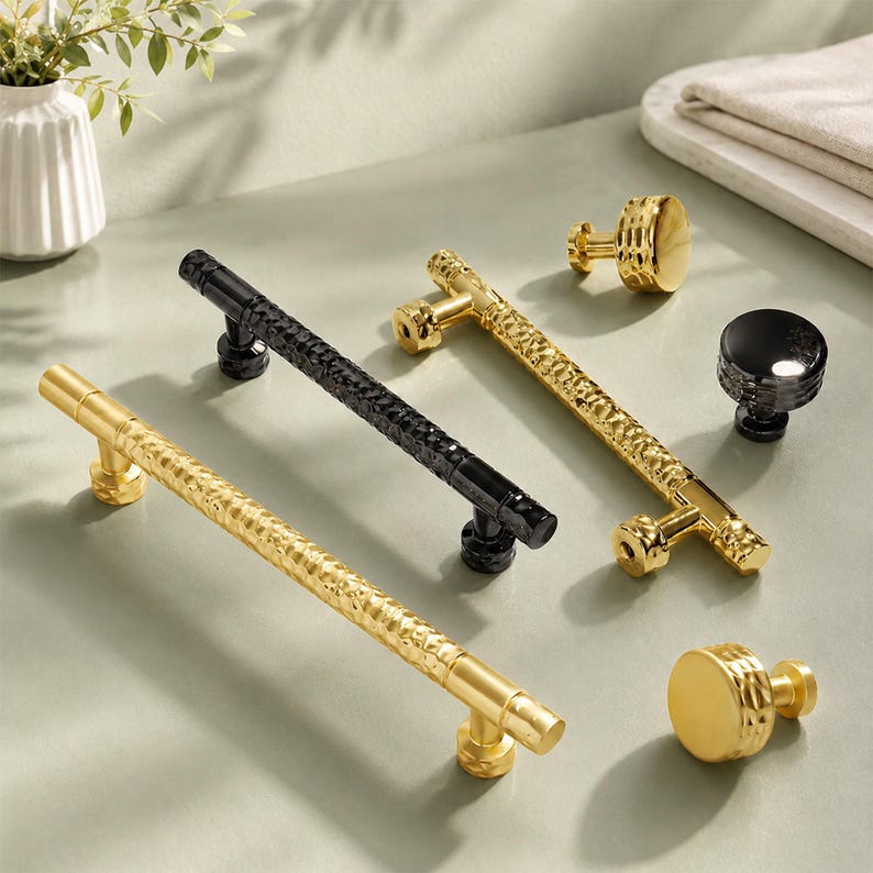 May include: A collection of cabinet hardware including two long bar pulls with a textured hammered finish in gold and black, one smooth gold bar pull, and three round knobs in gold, black, and textured gold, all featuring cylindrical mounting posts.