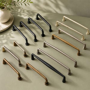 May include: A collection of metal cabinet pulls in various finishes including matte black, polished gold, brushed nickel, and antique brass. Each handle features a simple arched bar design with two circular mounting points at the ends.
