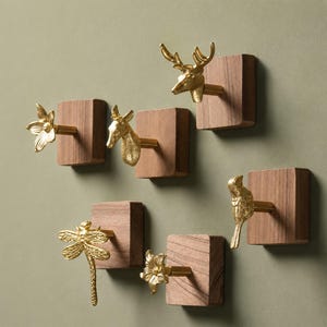 May include: A set of six wall hooks featuring gold-toned metal animal and floral figures mounted on square wooden blocks. The collection includes a deer head, a bird, a dragonfly, and three flower shapes, each attached to a natural wood base.