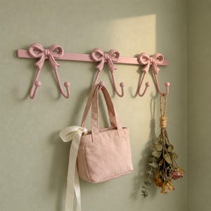 May include: A light pink wall-mounted coat rack featuring three decorative bow-shaped hooks. A small matching pink fabric handbag with a long white ribbon tie hangs from the center hook, and a bundle of dried flowers hangs from the right hook.