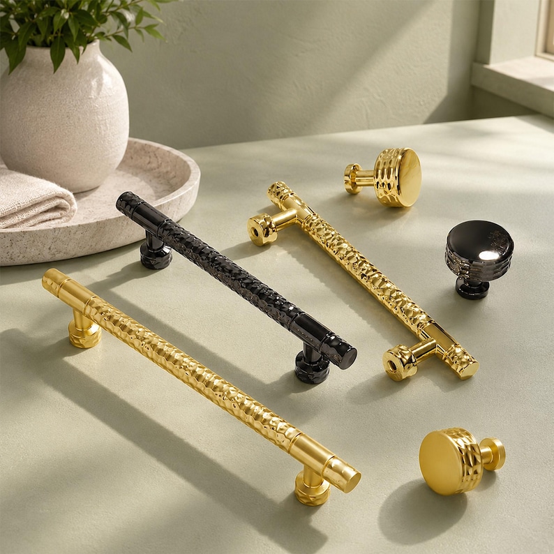 May include: A collection of cabinet hardware including two long, cylindrical drawer pulls with a hammered texture and two round knobs. One pull and one knob are finished in a polished gold tone, while the other pull and knob are finished in black.