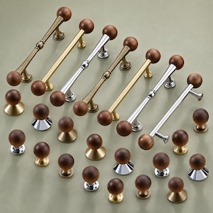 May include: A collection of cabinet hardware including several long bar pulls and numerous round knobs. The pieces feature a combination of natural wood spherical ends and metal bases in polished chrome, brushed brass, and antique bronze finishes.