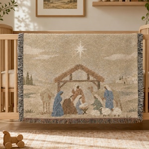 May include: A woven throw blanket featuring a beige and muted-tone nativity scene. The design depicts figures in blue and brown robes gathered under a wooden stable structure beneath a bright star, with camels and sheep in a desert landscape.