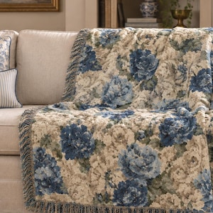 May include: A woven throw blanket featuring a blue and cream floral pattern with fringed edges, draped over a light-colored sofa. The textile displays detailed botanical motifs in shades of deep blue and off-white throughout the fabric.