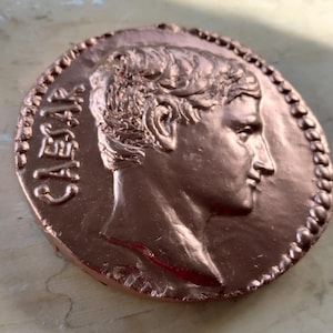 May include: A circular copper-colored medallion featuring a relief profile of a person facing right. The word CAESAR is embossed vertically on the left side. The edge of the medallion is decorated with a raised beaded border design.