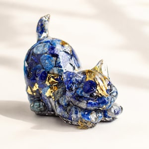Puede incluir: Una figura de gato hecha de pequeñas piedras de lapislázuli azul pulidas y copos metálicos dorados incrustados. La escultura representa un gato en posición agachada con la cola levantada, con un acabado de resina suave y brillante.