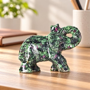 May include: A carved elephant figurine crafted from a composite of green zoisite and vibrant pink ruby mineral fragments. The sculpture features a polished, textured surface with a raised trunk and detailed ear, showcasing a natural speckled pattern.
