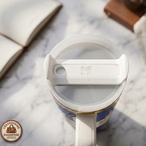 May include: A white plastic tumbler lid with a rotating closure mechanism featuring a circular straw opening and a small embossed winged creature logo. The word STRAW is molded into the top surface of the lid, which sits atop a blue vessel.
