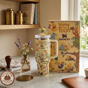 Peut inclure: Un gobelet Stanley jaune orné d'un motif de personnages Winnie l'ourson, doté d'une poignée blanche et d'un couvercle surmonté d'une petite figurine d'abeille. Le gobelet est présenté à côté de sa boîte en carton jaune assortie, décorée d'illustrations de personnages Winnie l'ourson et du logo Stanley.