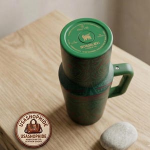 May include: A green Stanley brand tumbler with a handle and a patterned exterior featuring red and dark green geometric designs. The top lid displays the Stanley logo, a bear icon, and the text 40oz/1.18L. A circular logo patch with the text USASHOPHIDE is visible.
