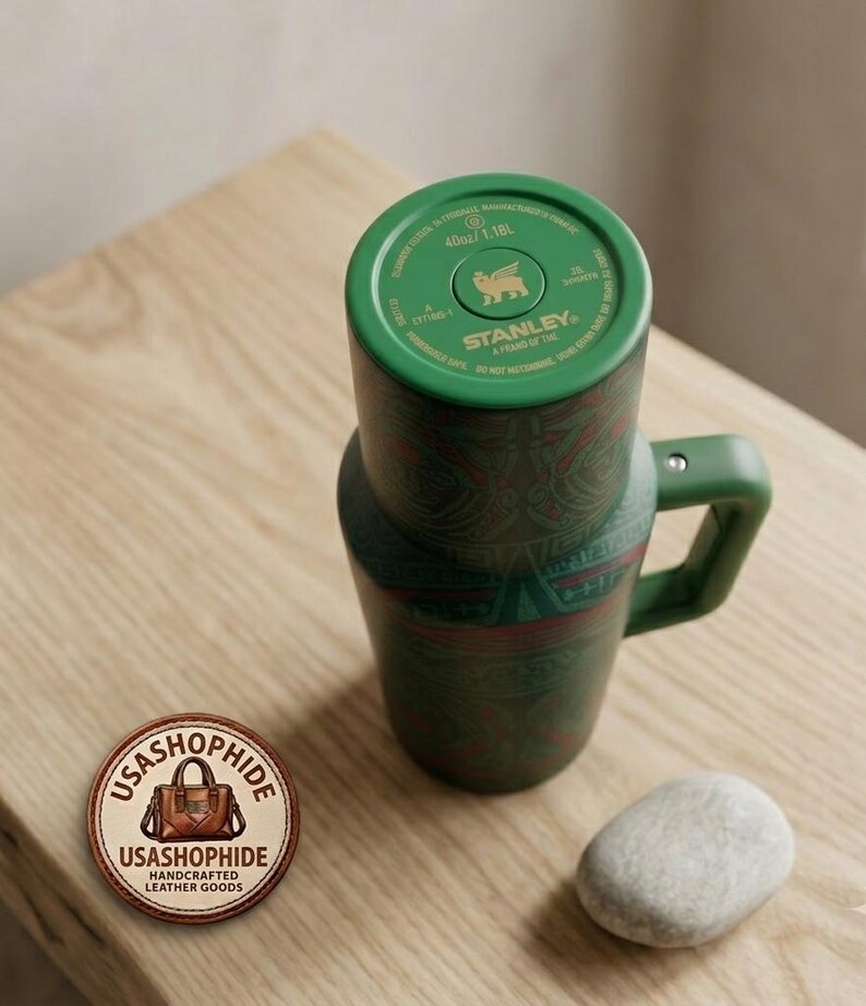 May include: A green Stanley brand tumbler with a handle and a patterned exterior featuring red and dark green geometric designs. The top lid displays the Stanley logo, a bear icon, and the text 40oz/1.18L. A circular logo patch with the text USASHOPHIDE is visible.