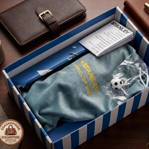 May include: A blue and white striped box containing a blue fabric pouch with the gold embroidered text STANLEY WORLD CUP 2026, a small white and black soccer ball charm in a plastic bag, and a white Stanley brand instruction booklet.