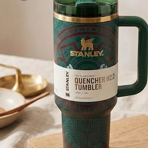 May include: A dark green Stanley Quencher H2.0 tumbler with a 40 oz capacity. The tumbler features a gold-colored rim, a matching green handle, and a translucent green lid. It is wrapped in a paper band labeled The Flowstate Quencher H2.0.