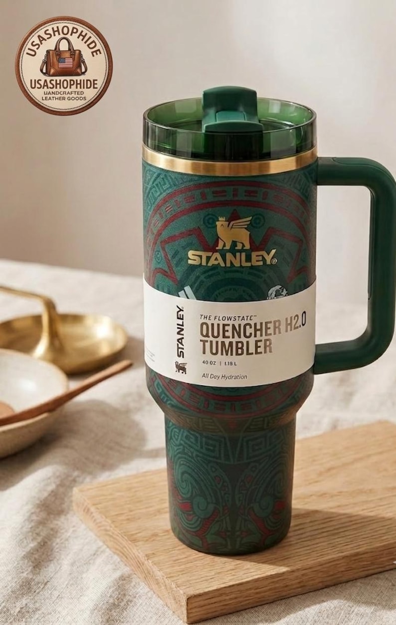 May include: A dark green Stanley Quencher H2.0 tumbler with a 40 oz capacity. The tumbler features a gold-colored rim, a matching green handle, and a translucent green lid. It is wrapped in a paper band labeled The Flowstate Quencher H2.0.