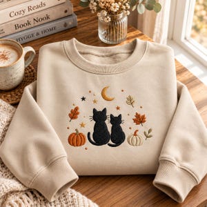 May include: A beige crewneck sweatshirt featuring an embroidered design of two black cats sitting side by side, surrounded by orange and white pumpkins, autumn leaves, small stars, and a crescent moon centered above the cats.